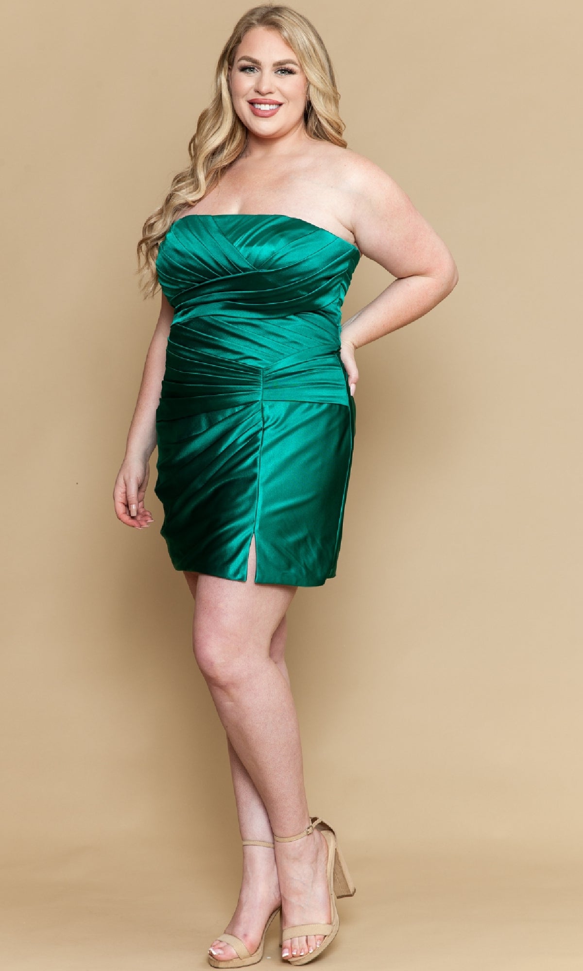 A woman poses in the Poly USA W1206 plus-size strapless party dress—an emerald green, ruched mini with a front slit—paired with beige high-heeled sandals, set against a beige background.